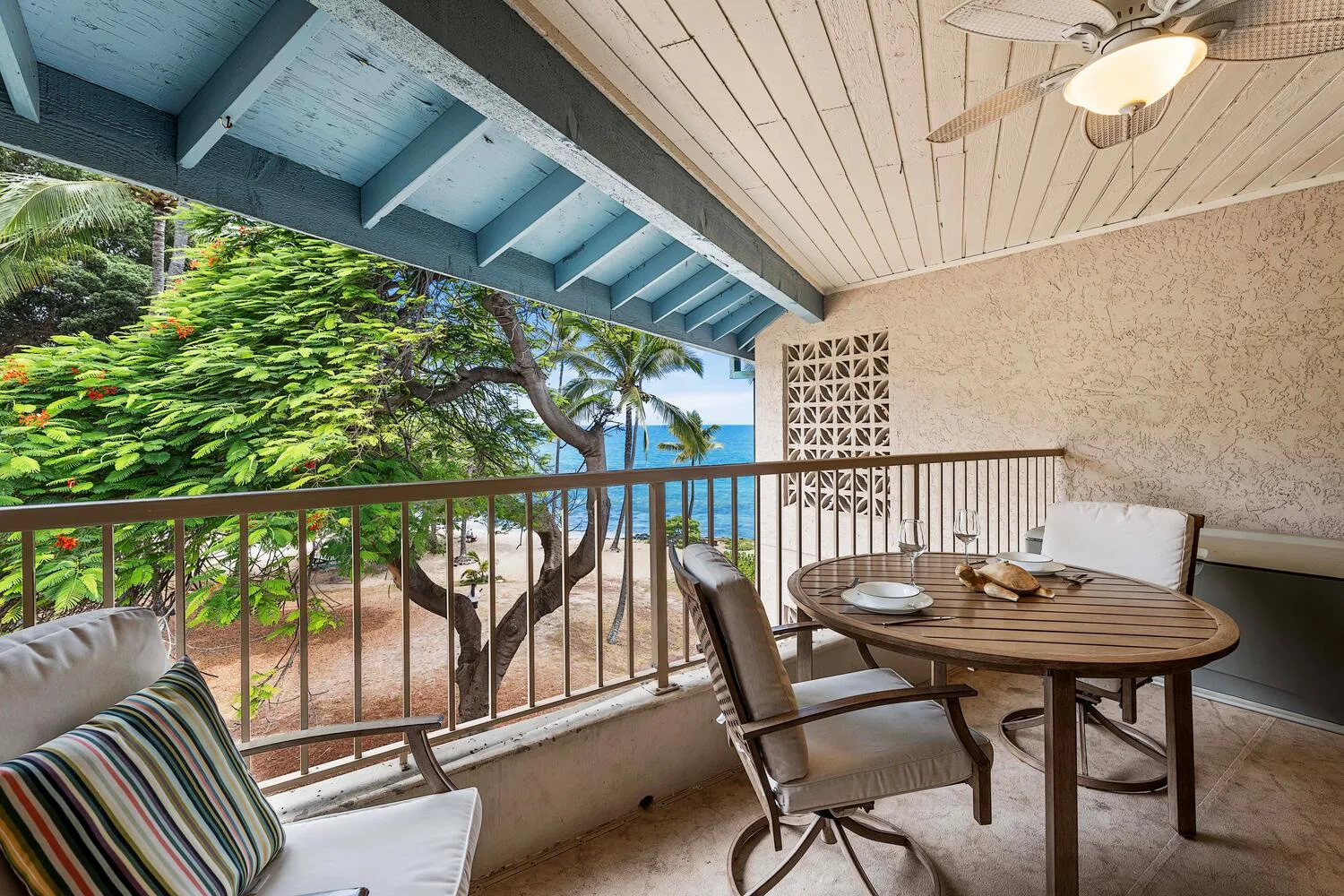 Un balcón cubierto con mesa de comedor y asientos da a la vegetación tropical y al mar.