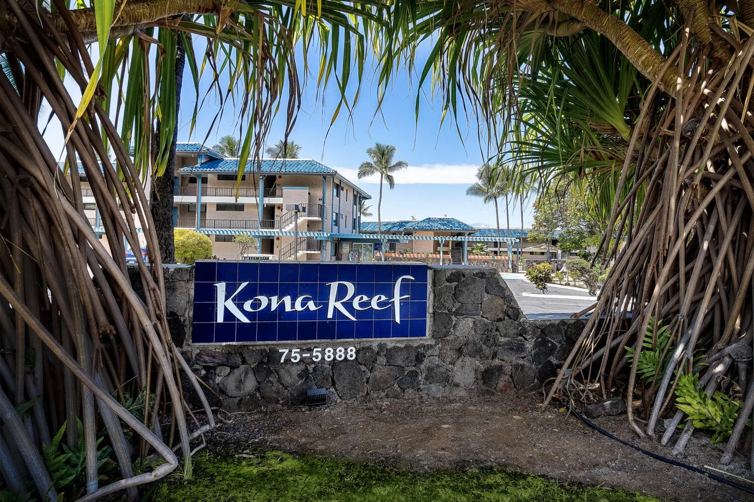 Un muro de piedra de entrada muestra un letrero azul de Kona Reef bajo follaje tropical.