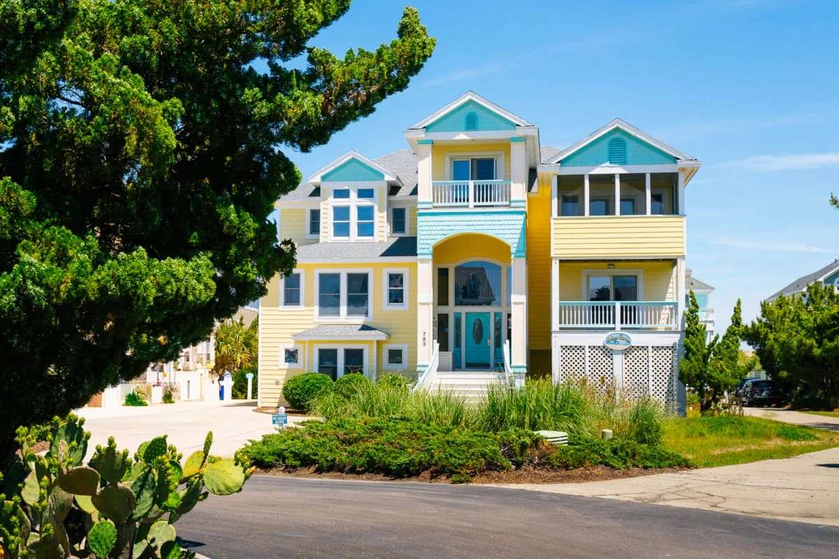 A large yellow house with turquoise trim stands beside a tree-lined street.