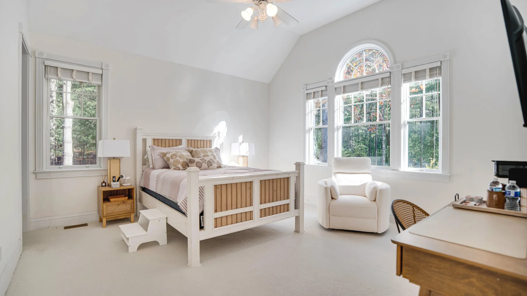 A bright bedroom with a white bed, armchair, and large windows.