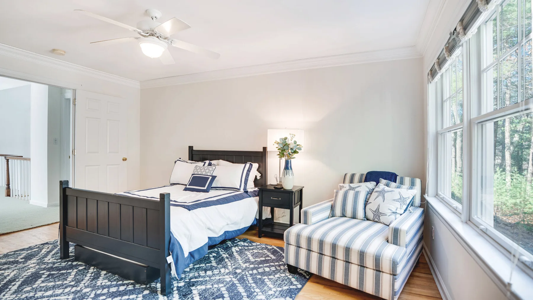 A bright bedroom with a bed, an upholstered armchair, and large windows.
