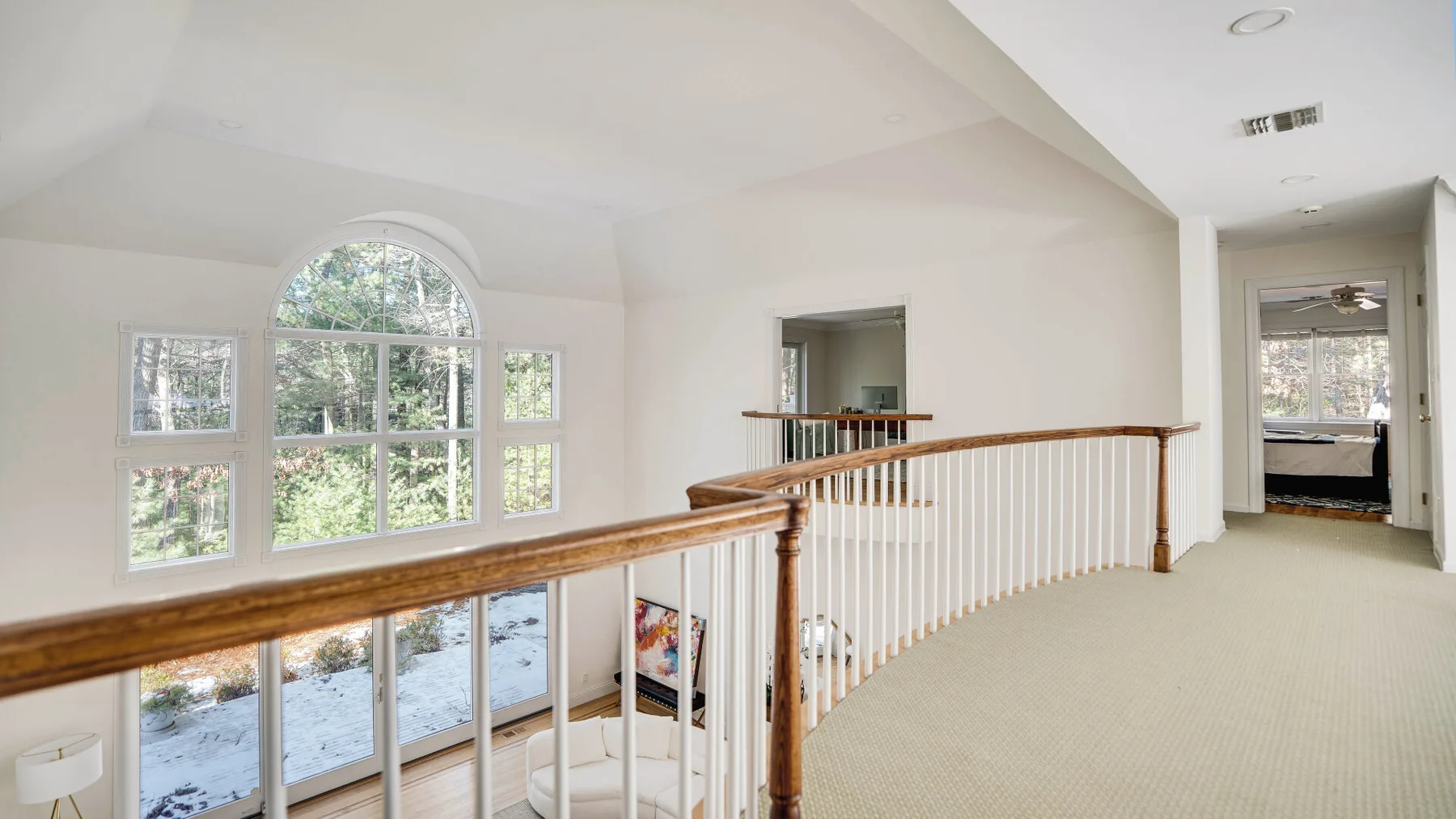 An upstairs landing with a wooden railing overlooks a bright room with large windows.