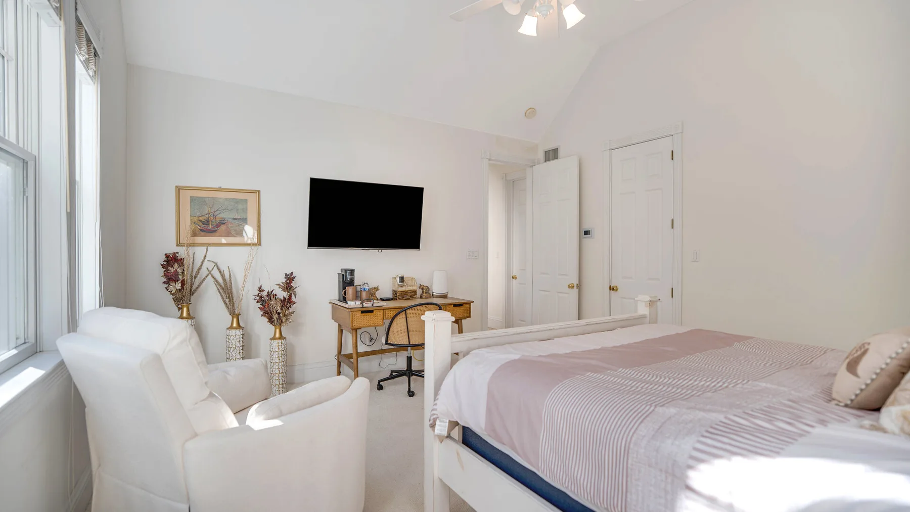 A bright bedroom with a bed, seating, and a desk beneath a wall-mounted television.