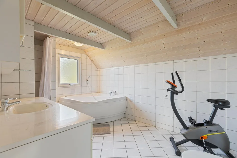 Un baño alicatado con bañera, lavabo y máquina de ejercicio bajo un techo inclinado de madera.