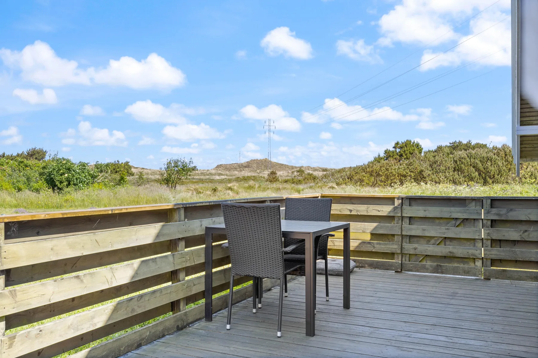 Una terraza de madera con asientos al aire libre da a dunas cubiertas de hierba bajo un cielo azul.