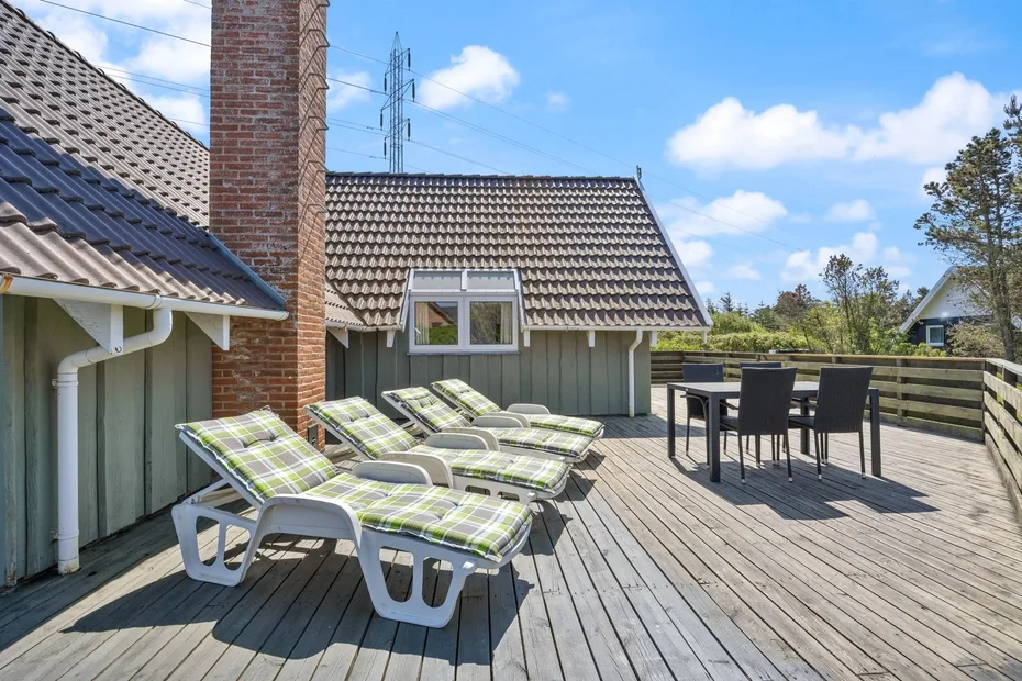Una terraza de madera tiene asientos reclinables y una mesa de comedor al aire libre bajo un cielo azul.
