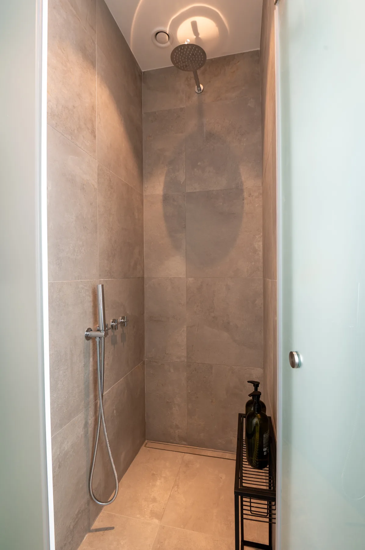A tiled shower area with a handheld showerhead and dark bottle on a small stand.