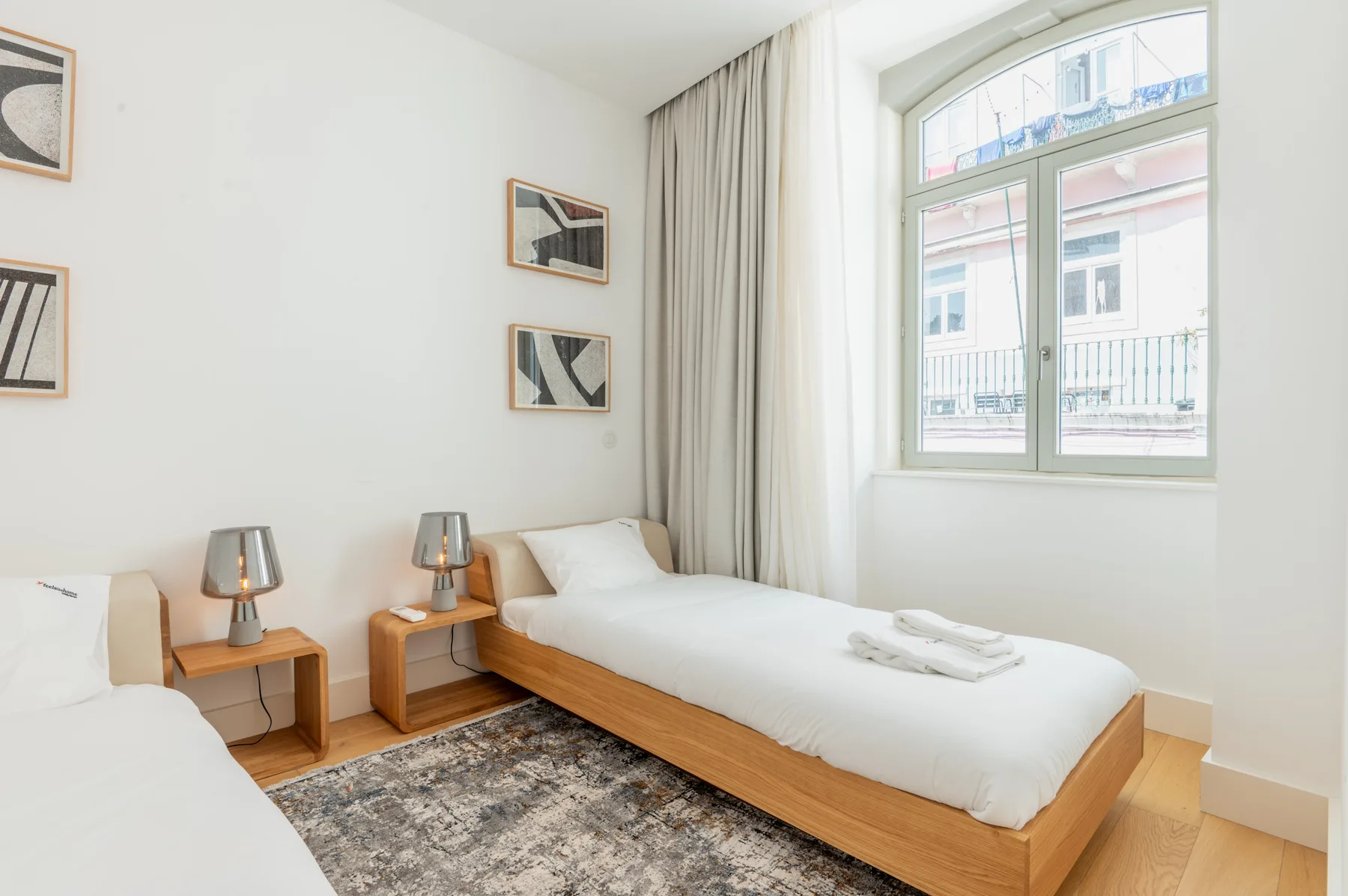 A bright bedroom with two single beds, bedside tables, and a large window.
