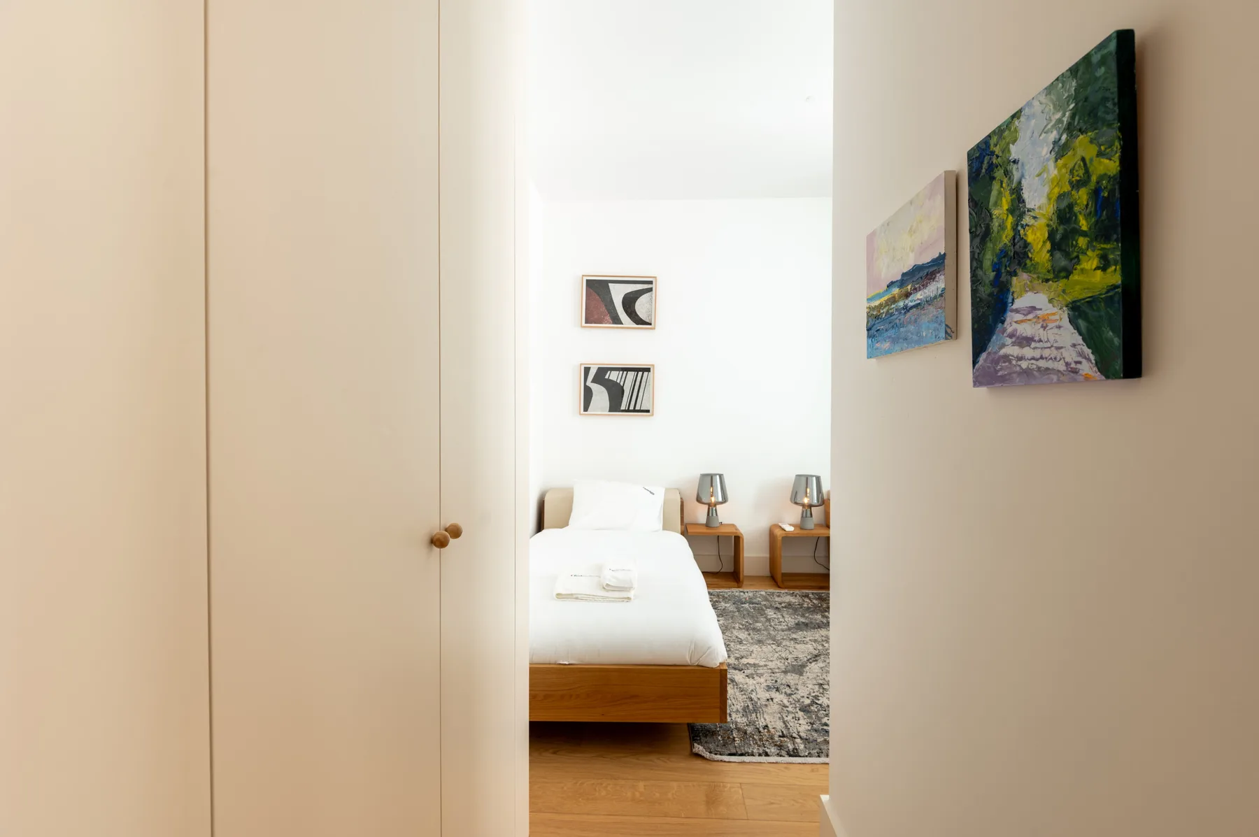 A bedroom with a bed, bedside tables, and artwork visible through an open doorway.