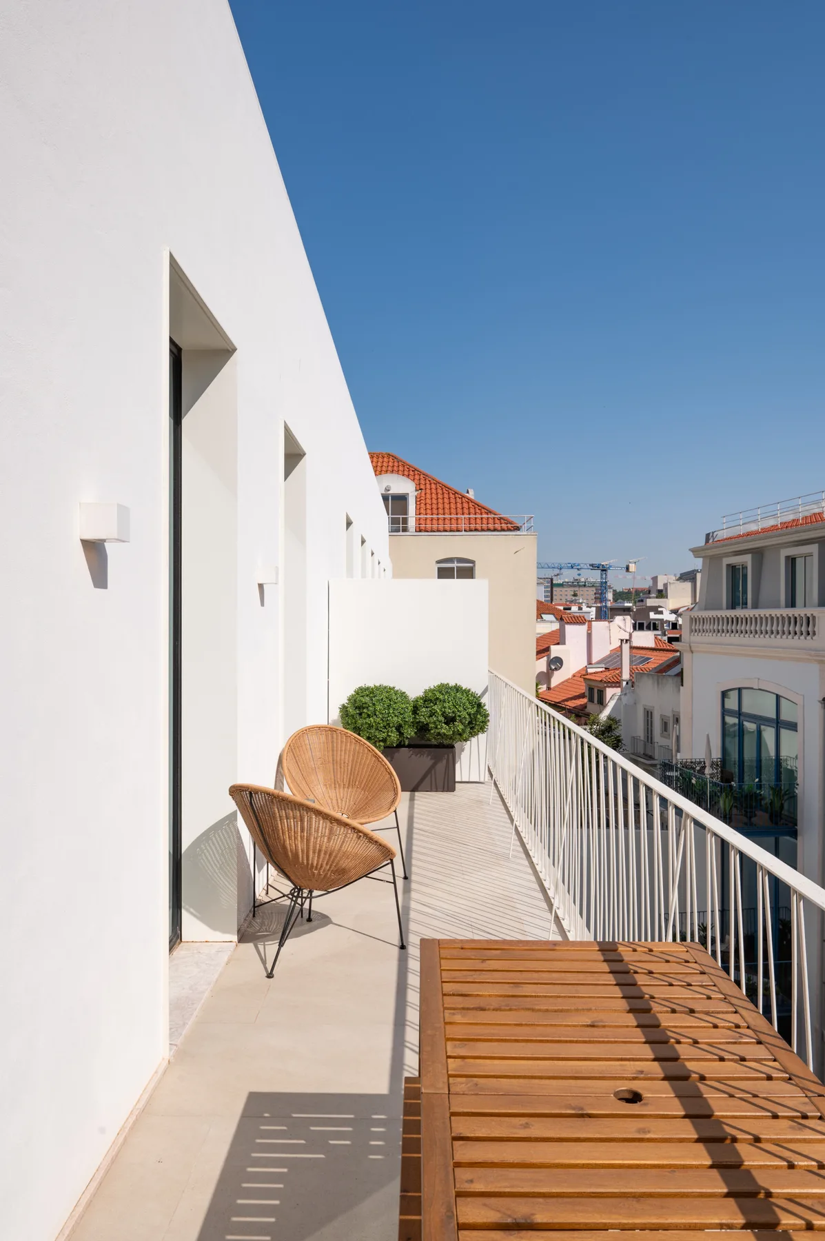 A sunlit balcony with woven seating, a potted shrub, and a wooden table.
