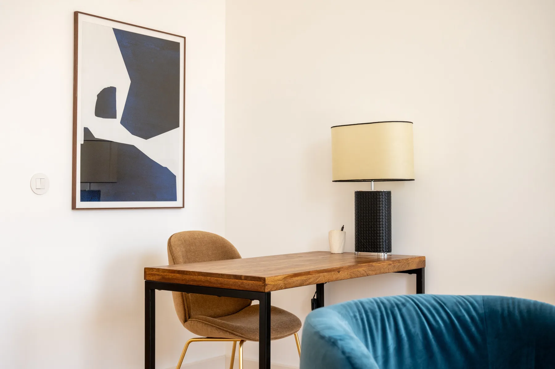 A desk and chair sit against a light wall, with a lamp and framed artwork above.