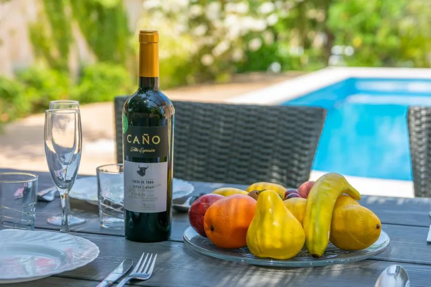 Una mesa de comedor en un patio con fruta y vino junto a una piscina.