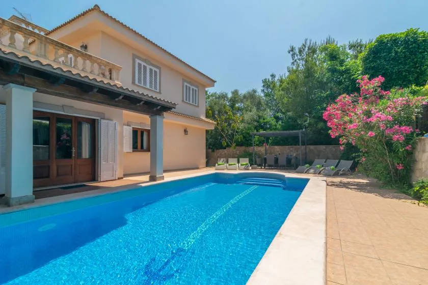 Una piscina junto a una casa, con asientos de descanso y árboles en flor cerca.