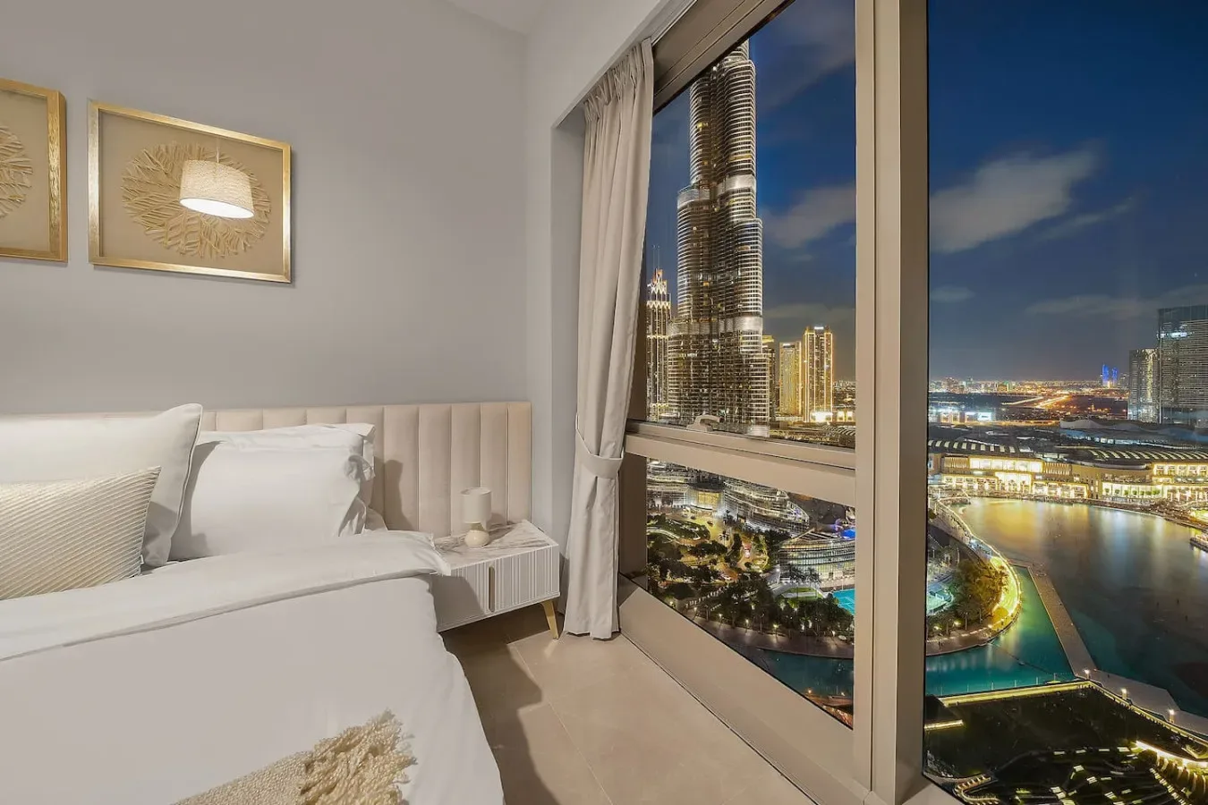 Un dormitorio con una cama bien hecha junto a una gran ventana con vistas a la ciudad de noche.