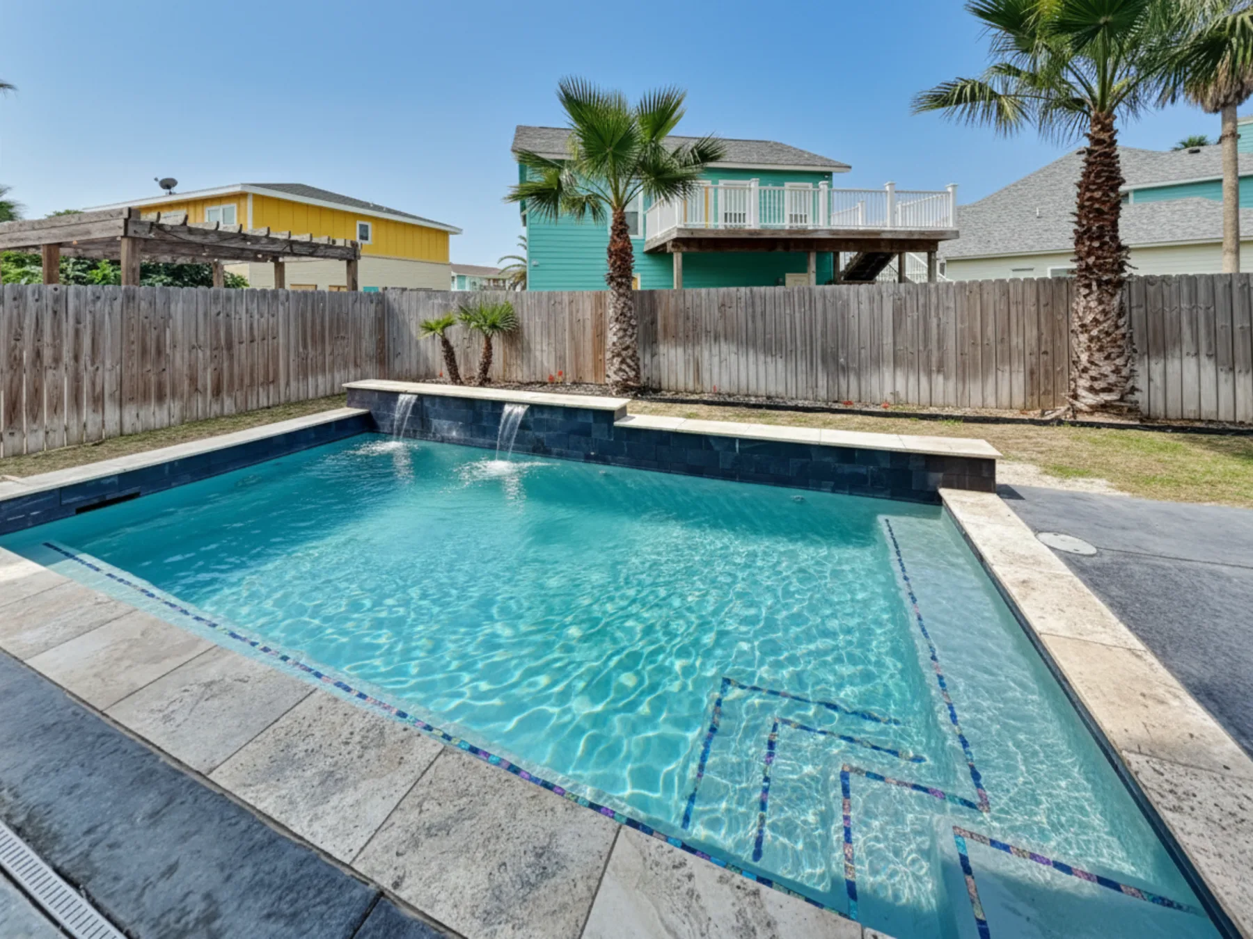 A backyard swimming pool sits beside a wooden fence and palm trees.