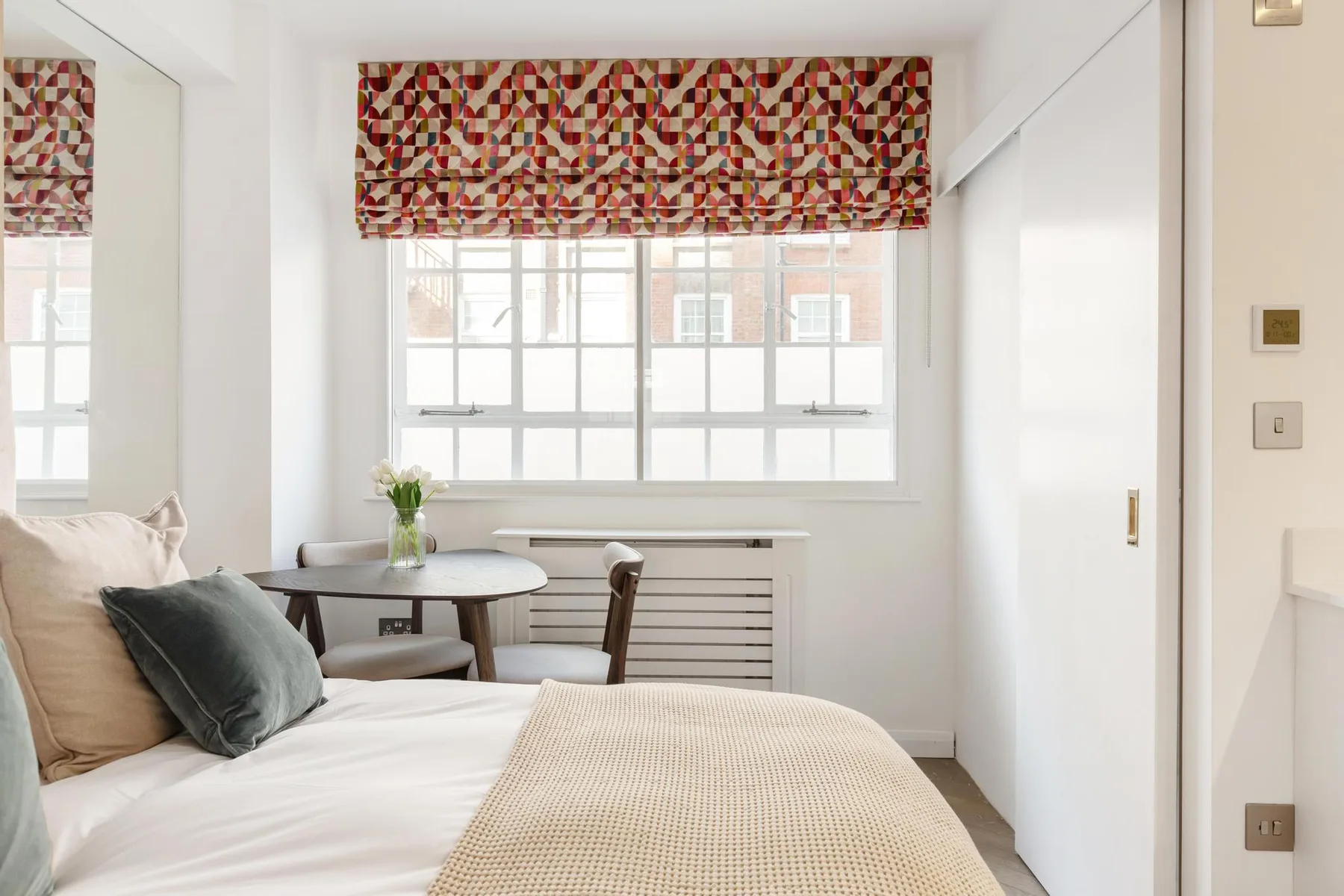 Un dormitorio luminoso con una cama junto a una ventana grande y una persiana estampada.
