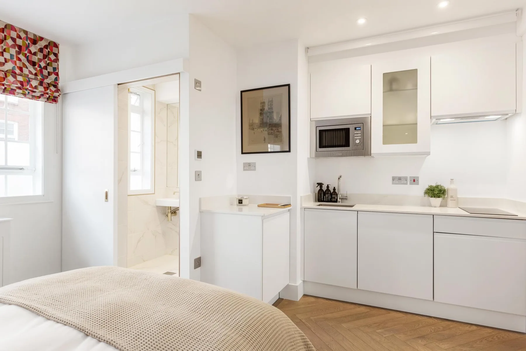 Una pequeña cocina blanca y luminosa con microondas y una cama en primer plano.