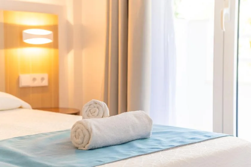 A bedroom with neatly rolled towels on the bed beside a bright window.