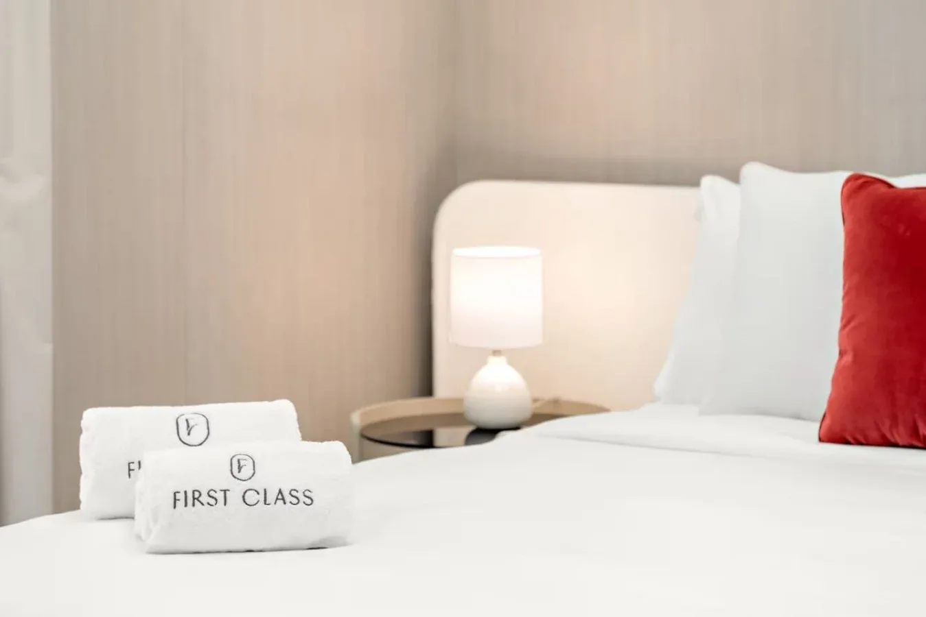Un dormitorio con ropa de cama blanca, una lámpara de noche y un cojín rojo.