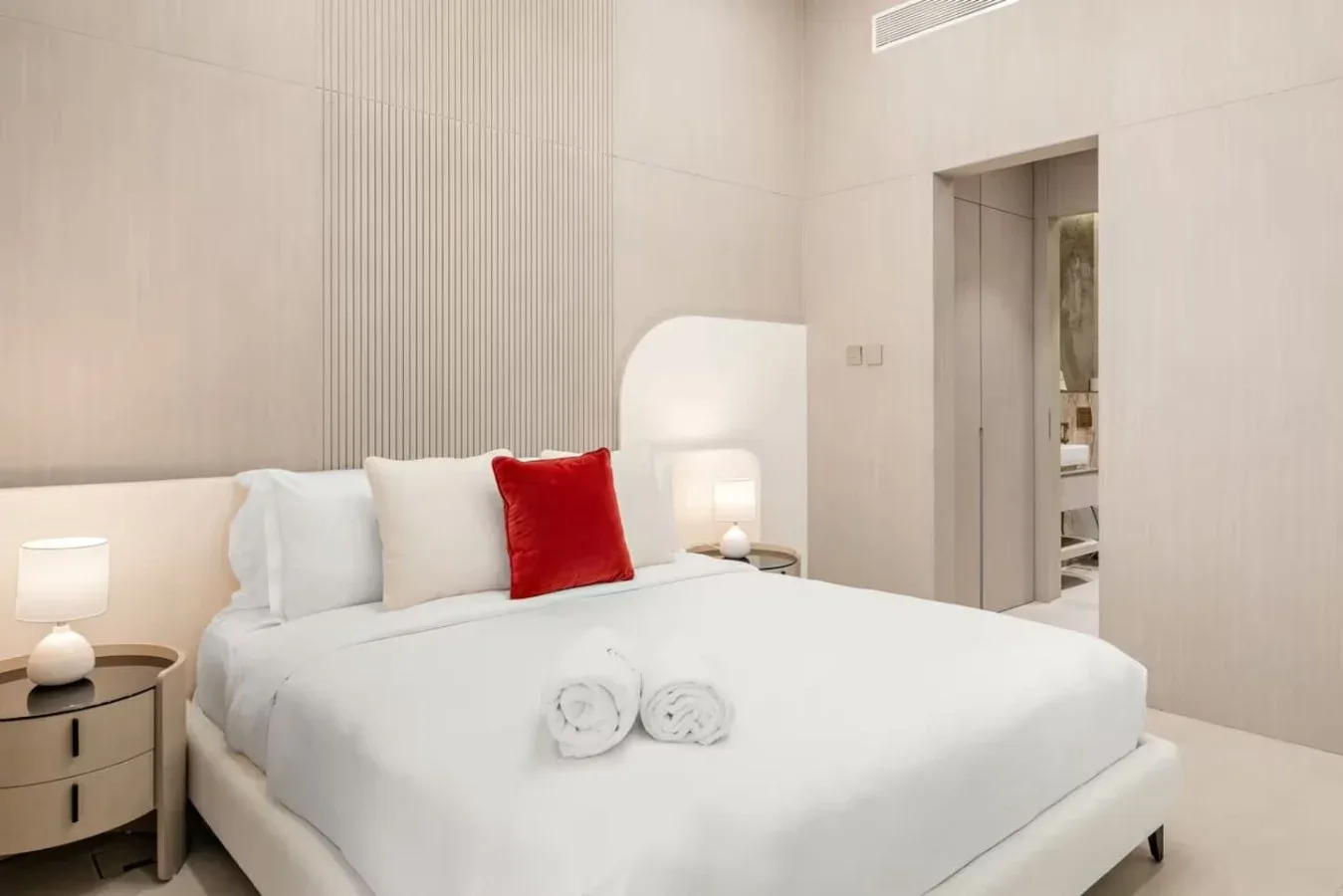 Un dormitorio luminoso con una cama tendida y un cojín rojo.
