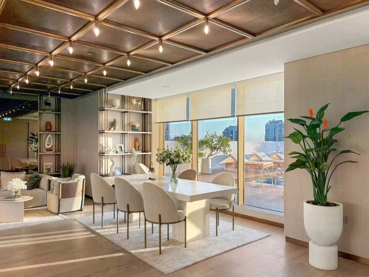 Un comedor luminoso tiene una mesa larga junto a grandes ventanas y plantas en macetas.