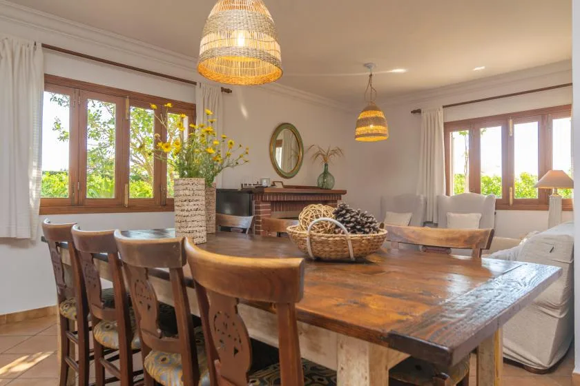 A dining area with a large wooden table, chairs, and windows overlooking greenery.