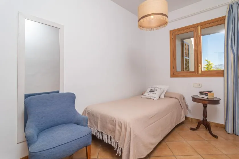 A bedroom with a single bed, a blue armchair, and a window.