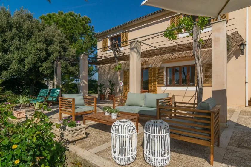 A shaded outdoor seating area sits beside a house, with a wooden pergola overhead.
