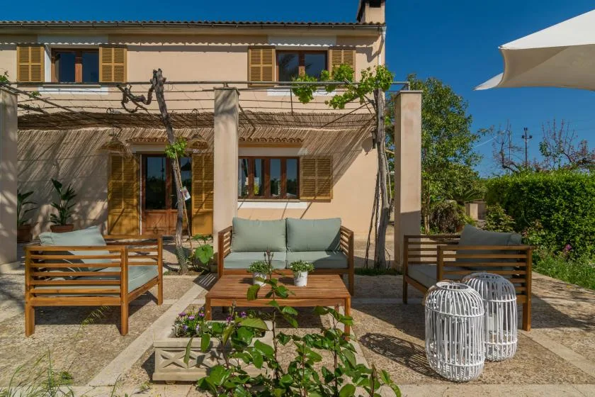 A sunlit outdoor patio has seating beneath a wooden pergola beside a house.