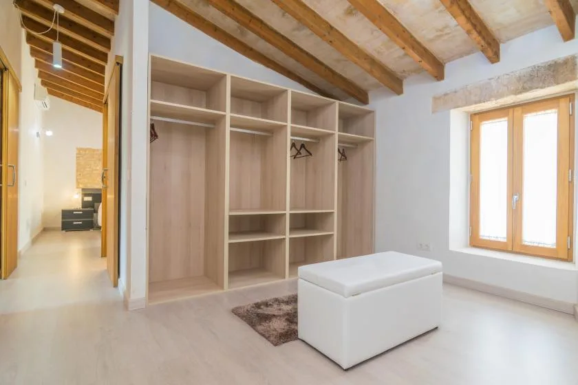 Una habitación luminosa tiene estanterías abiertas de madera, un otomano blanco y una ventana.