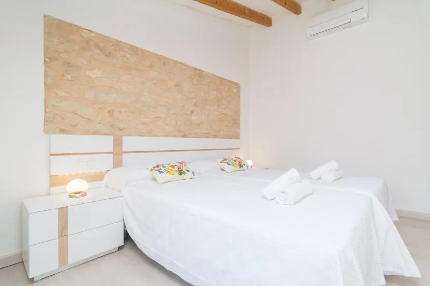 Un dormitorio luminoso con dos camas vestidas de blanco y una pared decorativa de piedra clara.