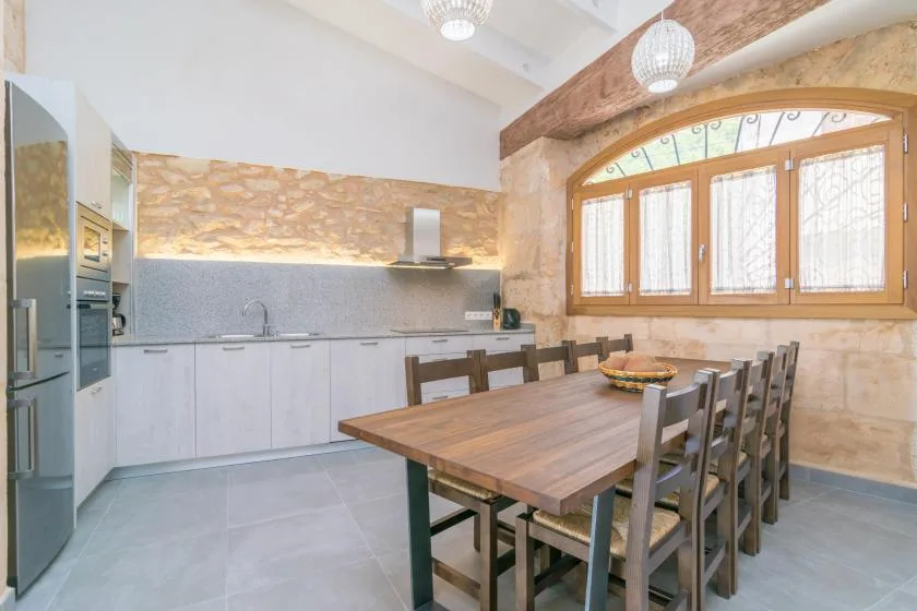 Una amplia cocina con zona de comedor, una larga mesa de madera y grandes ventanas.