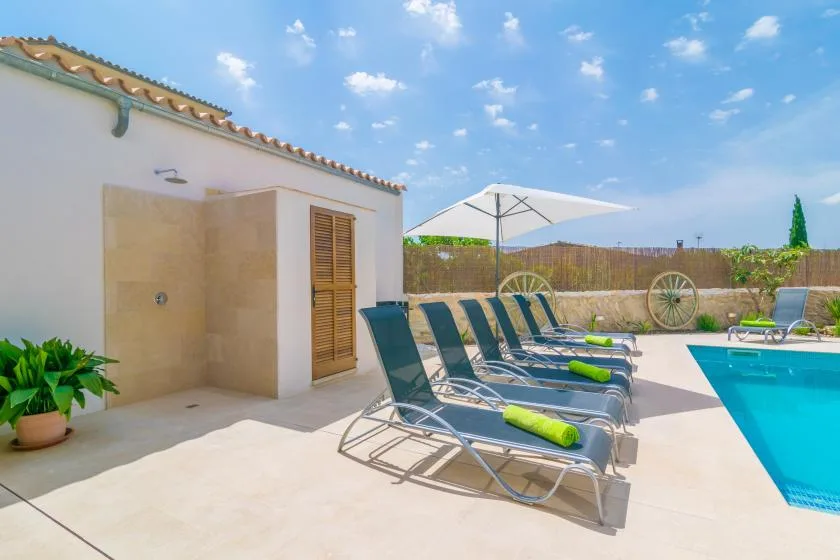 Un patio soleado junto a la piscina, con asientos, una sombrilla y una ducha exterior.