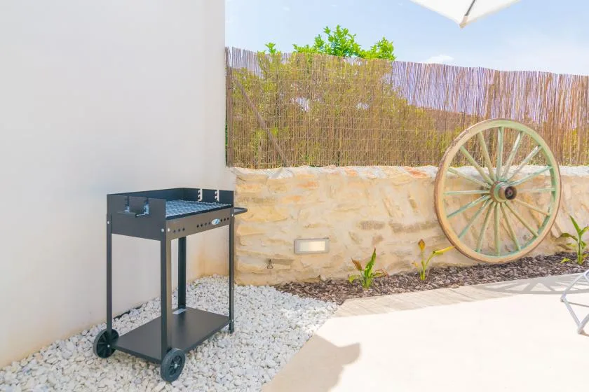 Un patio exterior con una parrilla metálica junto a un muro de piedra y una rueda decorativa.