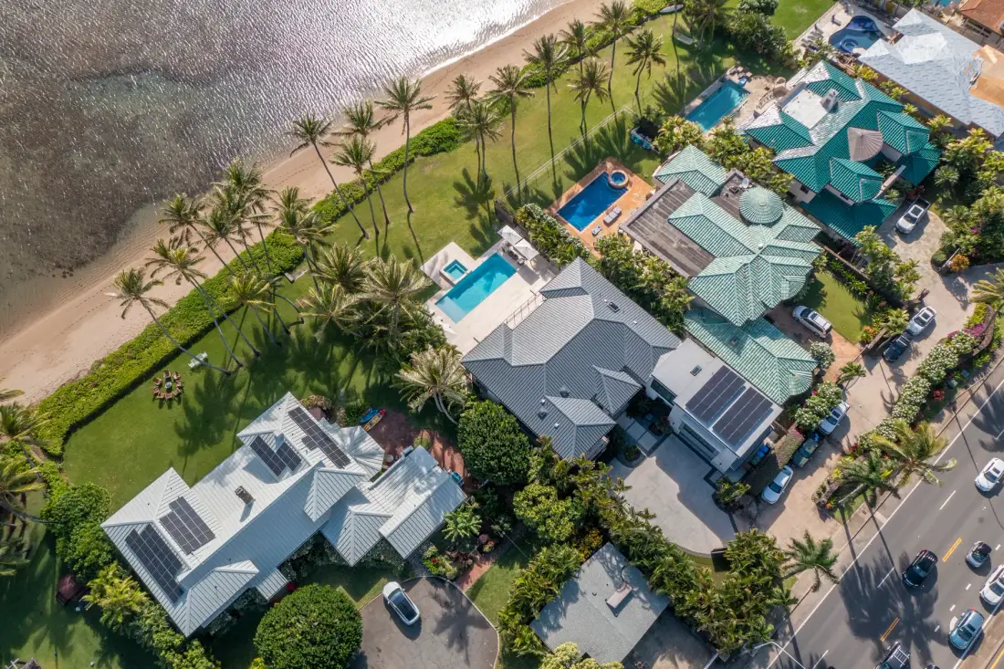 Una vista aérea muestra casas costeras, palmeras, piscinas y una playa.