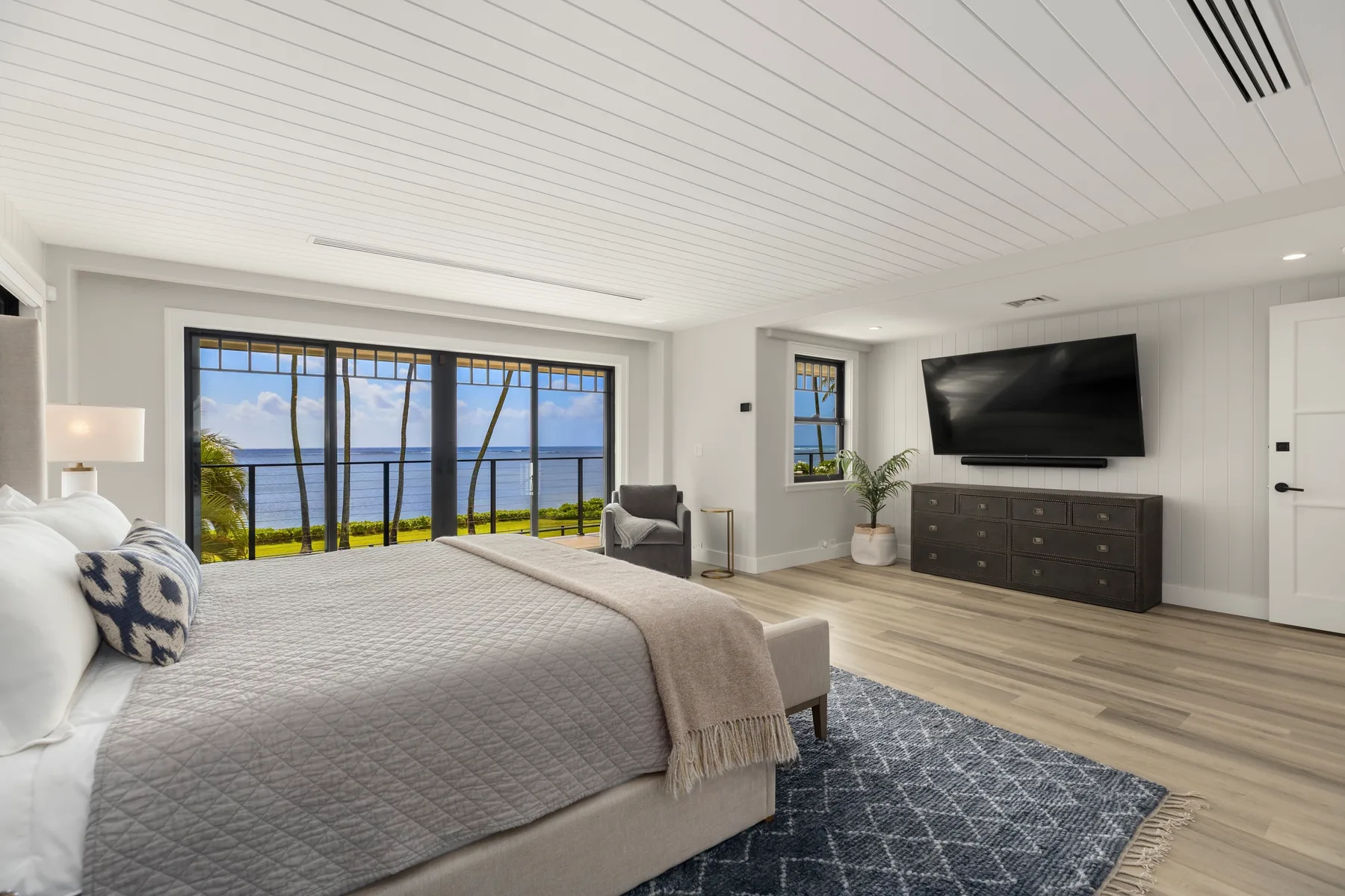 Un dormitorio luminoso con una cama grande, un televisor en la pared y ventanas con vistas al agua.