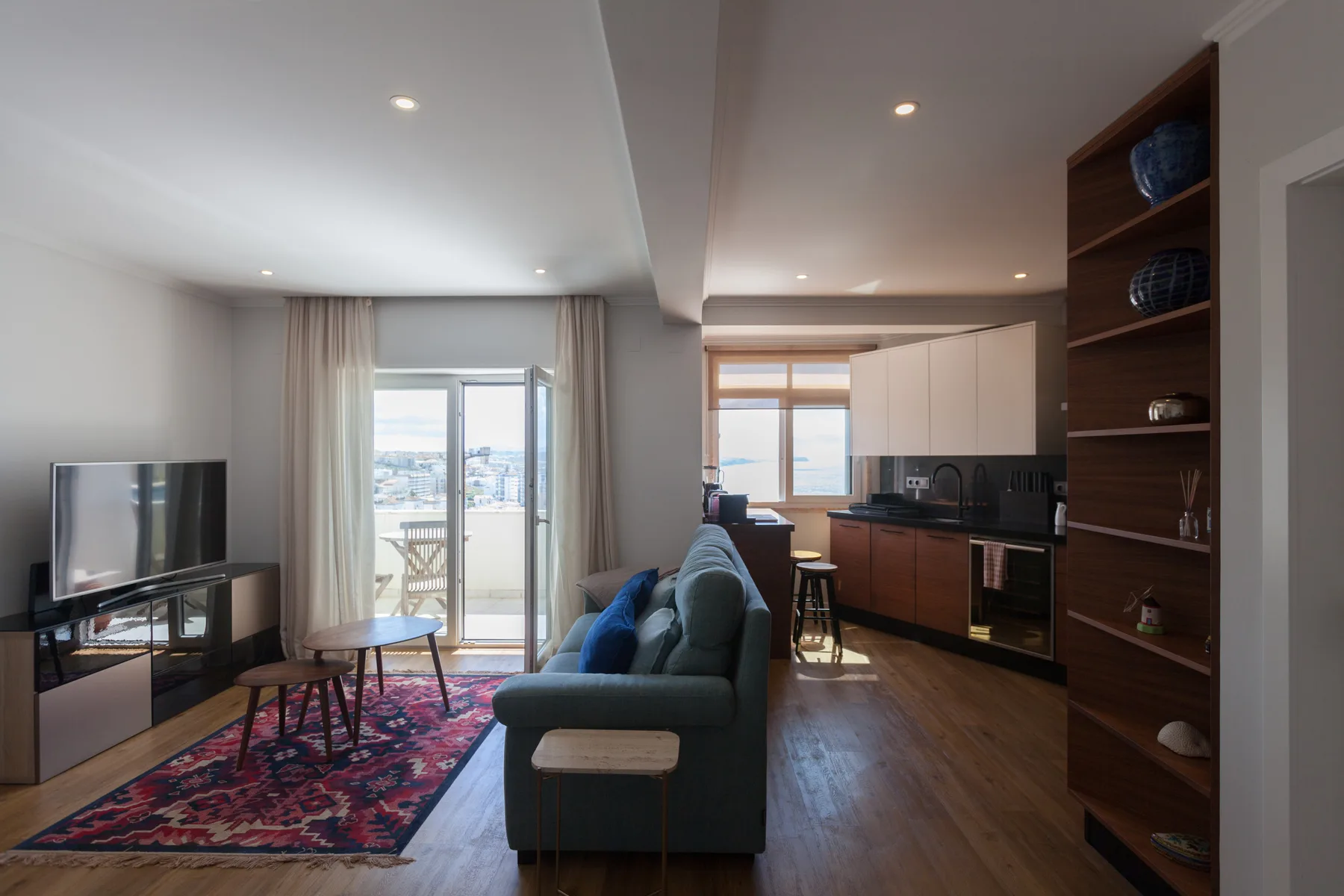 Una sala de estar conecta con una cocina, con asientos frente al televisor y puertas al balcón.