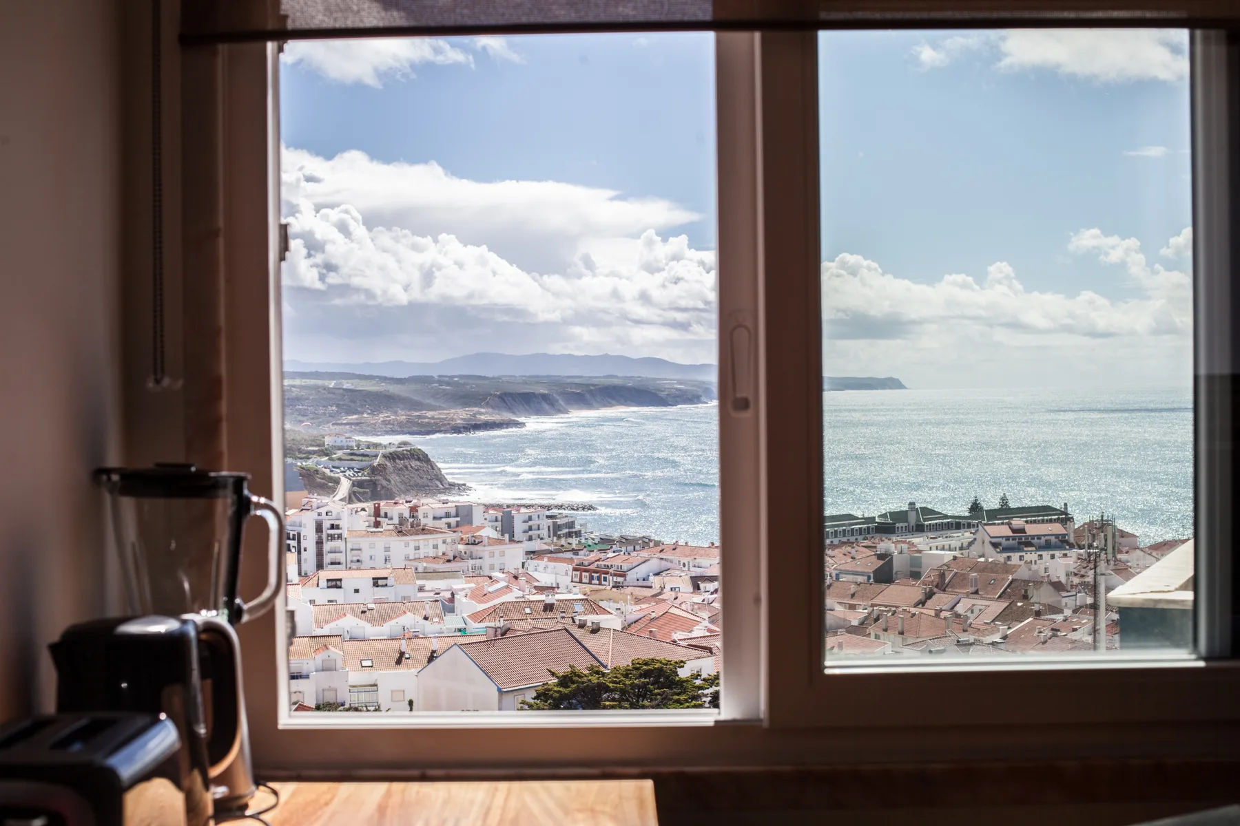 Una ventana enmarca un pueblo costero y el océano al fondo.