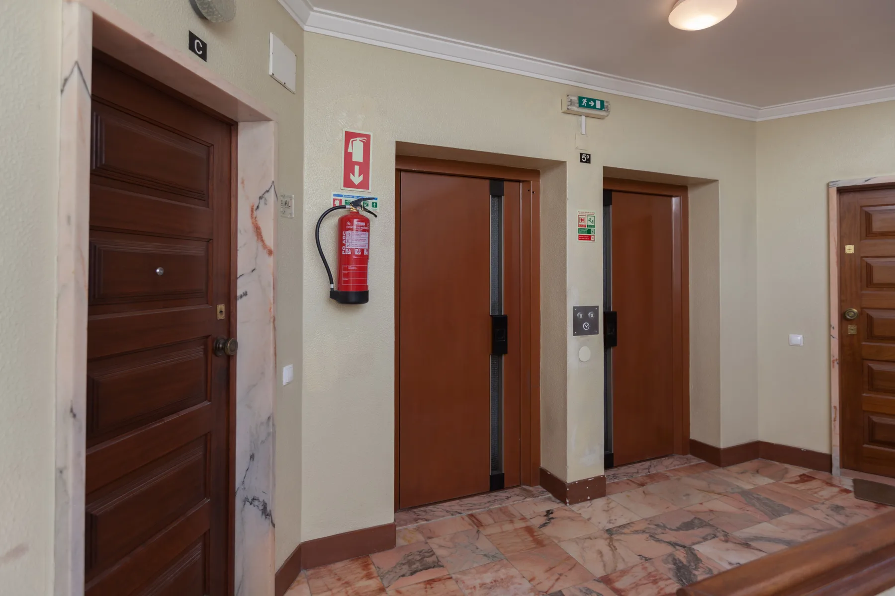 Un pasillo interior con puertas de madera, puertas de ascensor y un extintor.