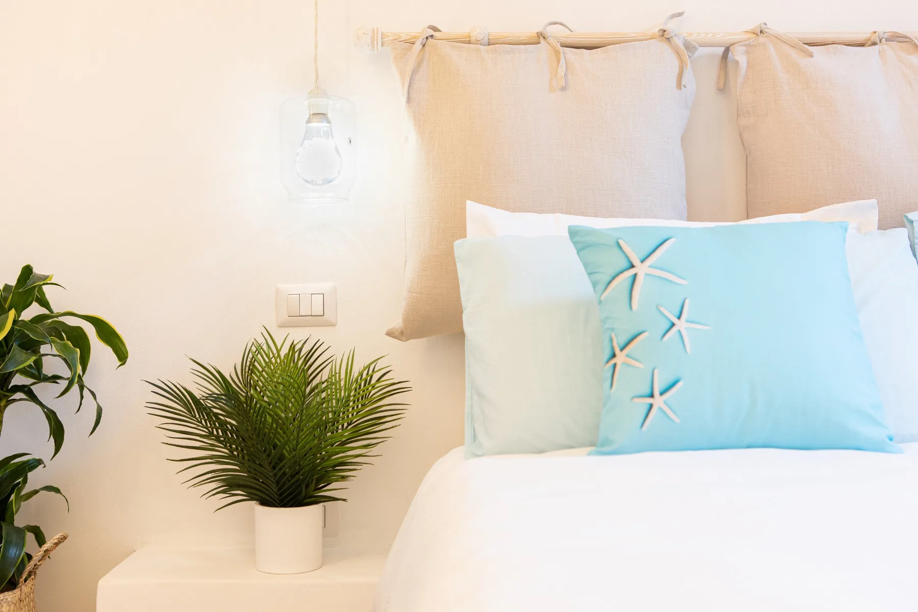 A bright bedroom features a bed with pale blue pillows and green plants.