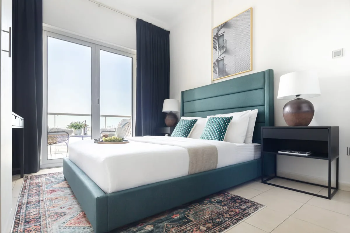 Un dormitorio con una cama verde azulado, ropa de cama blanca y grandes puertas de vidrio.