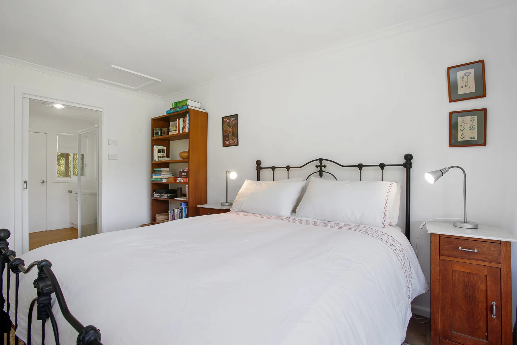 A bright bedroom with a large bed, bedside tables, and a wooden bookcase.