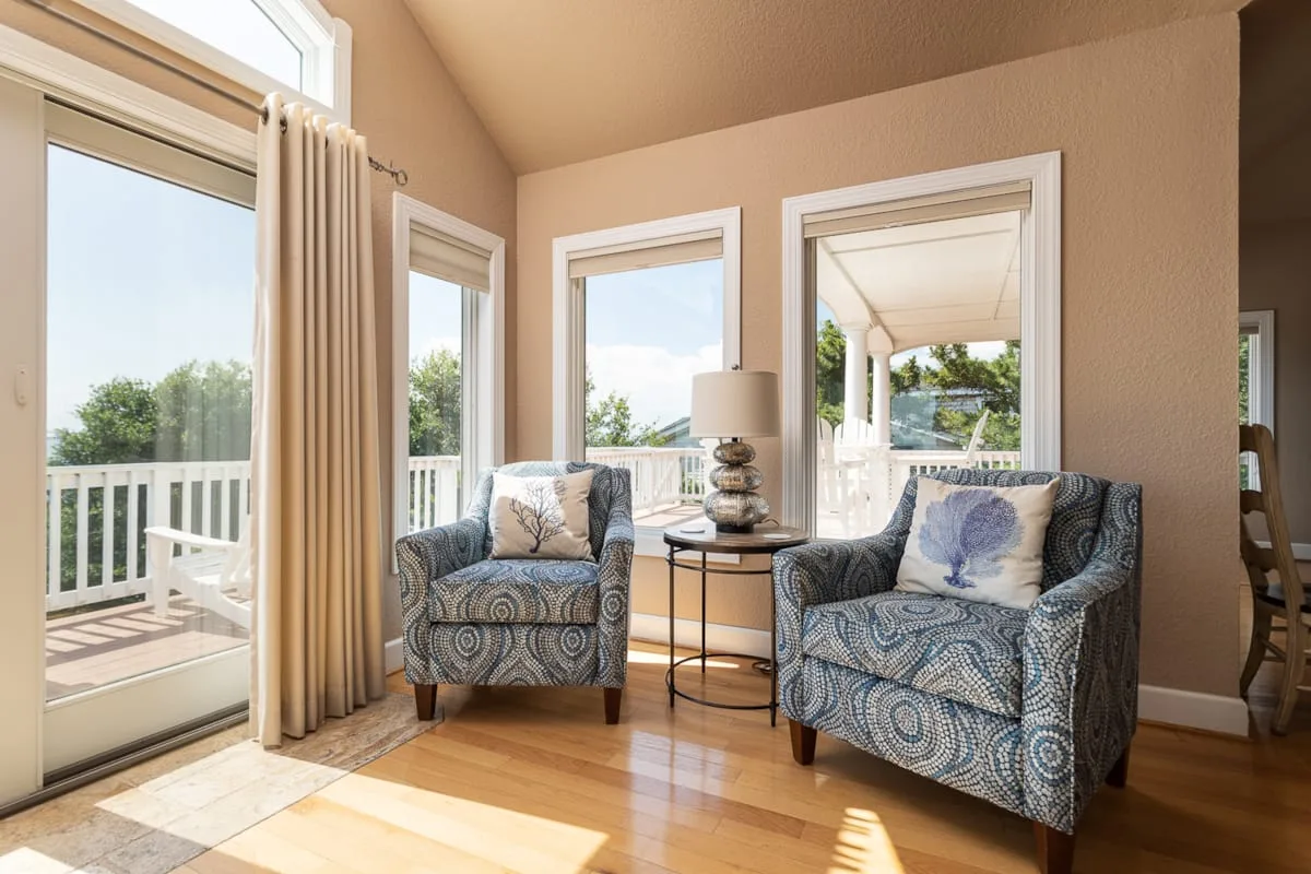 Una zona de estar con dos sillones estampados junto a ventanas luminosas y una puerta de vidrio.