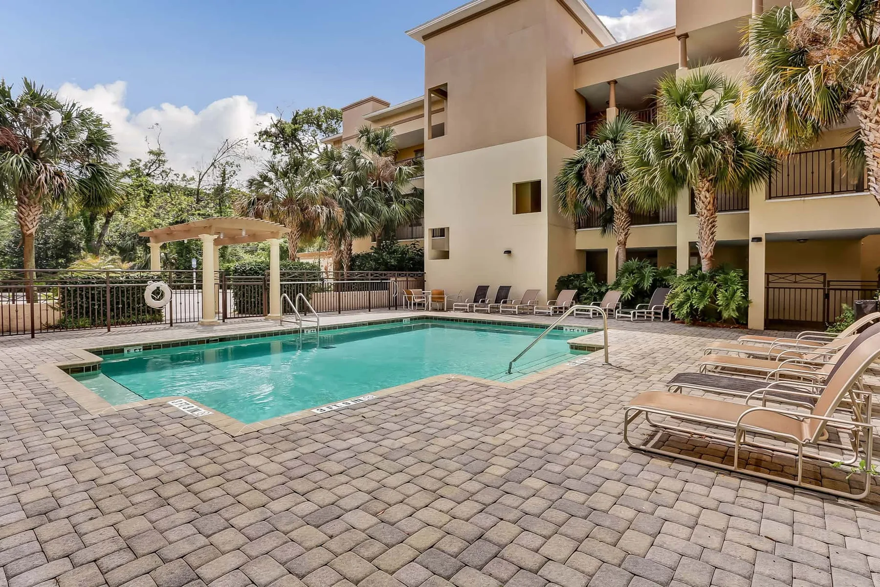 An outdoor swimming pool is surrounded by paved decking, lounge chairs, and palm trees.