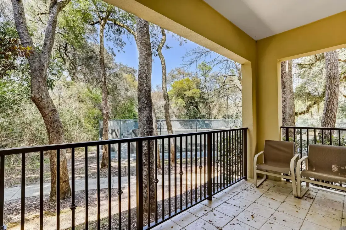 A covered balcony overlooks trees, with seating beside a black railing.