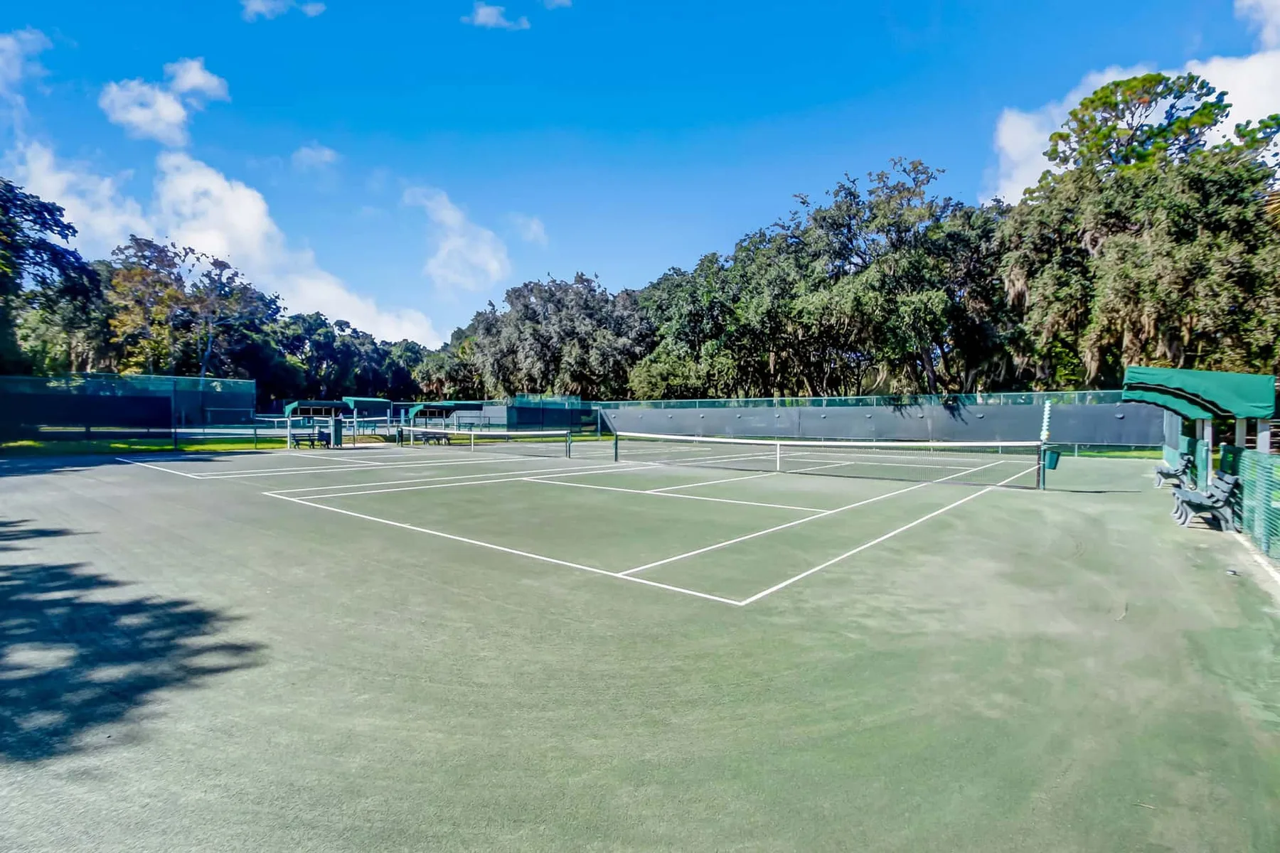 Outdoor tennis courts are surrounded by leafy trees and green fencing.