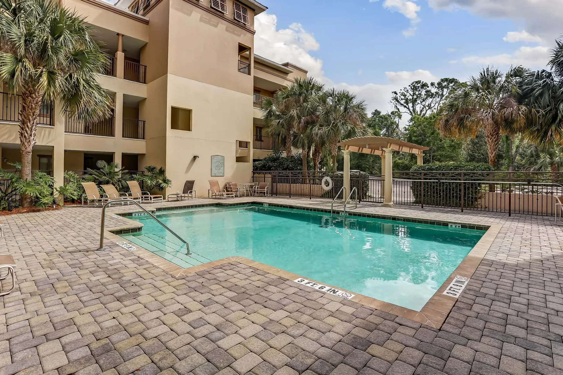 An outdoor swimming pool beside a multi-story building, with palm trees and paved decking.