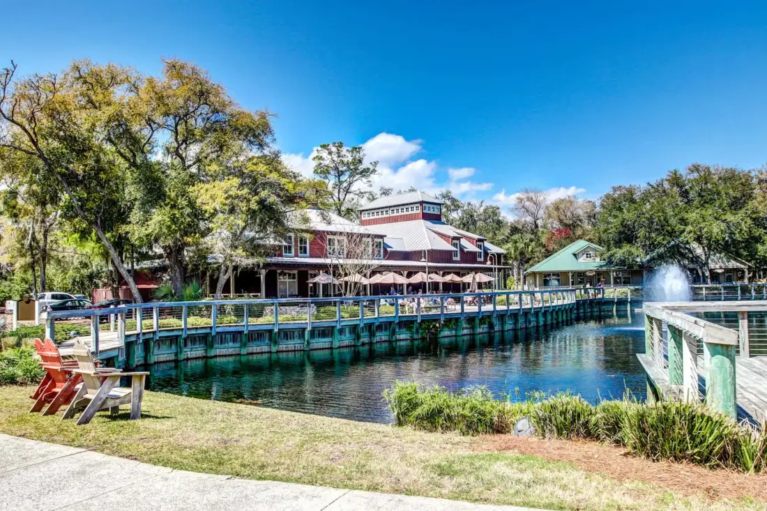 A waterside restaurant and covered walkway stand beside a pond with a fountain.