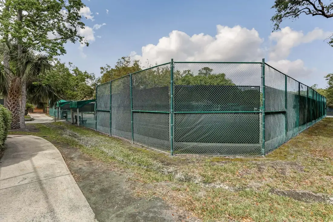 An outdoor fenced court sits beside a paved walkway and trees.