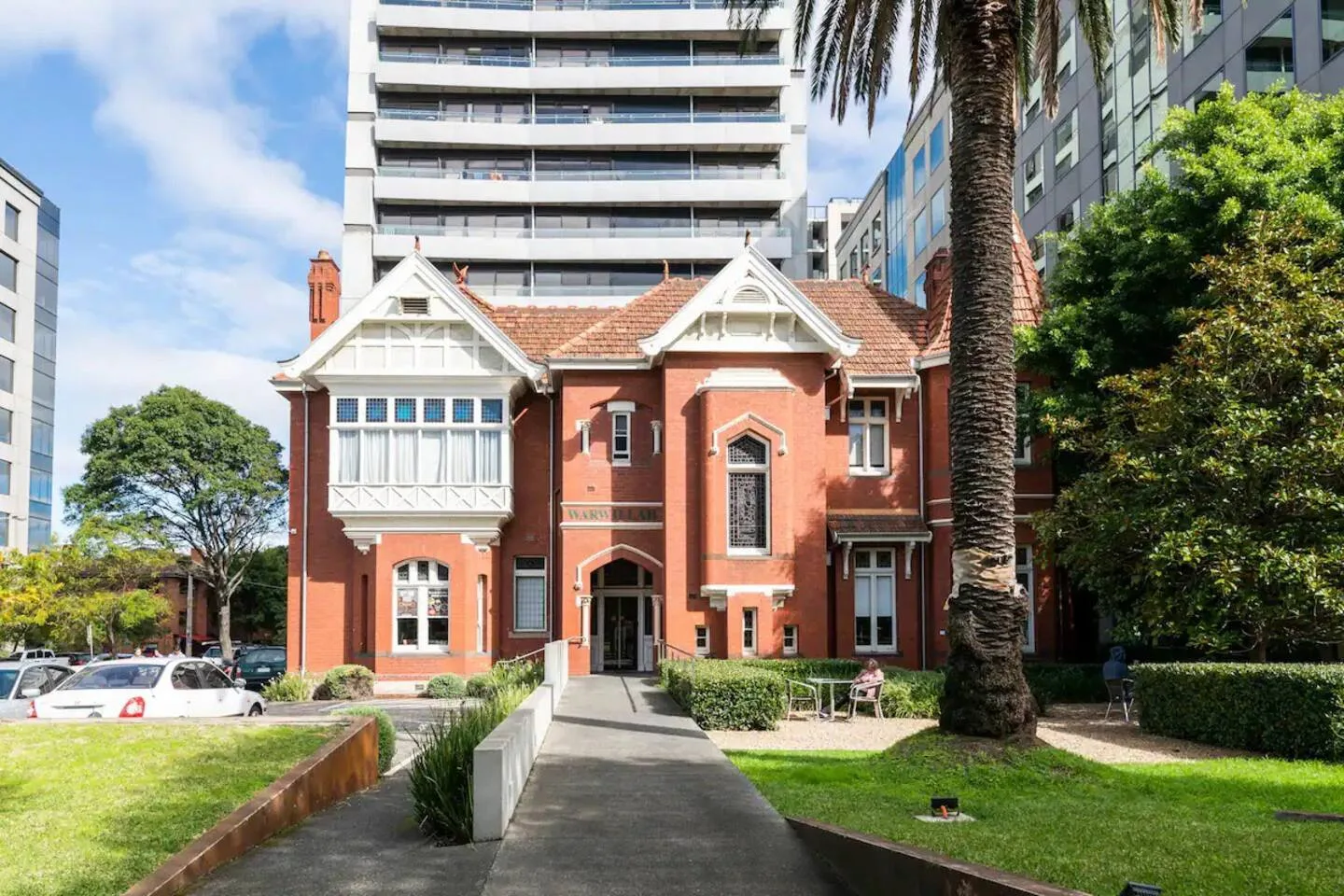 A red heritage-style building stands beside a paved walkway, with modern buildings behind it.